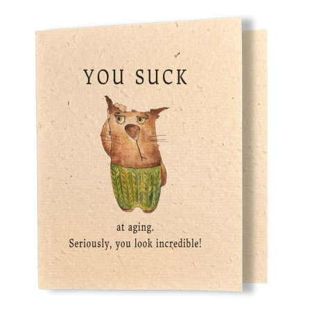A kraft seed paper birthday card featuring a grumpy watercolour cat in a green sweater with the text "YOU SUCK at aging. Seriously, you look incredible!"
