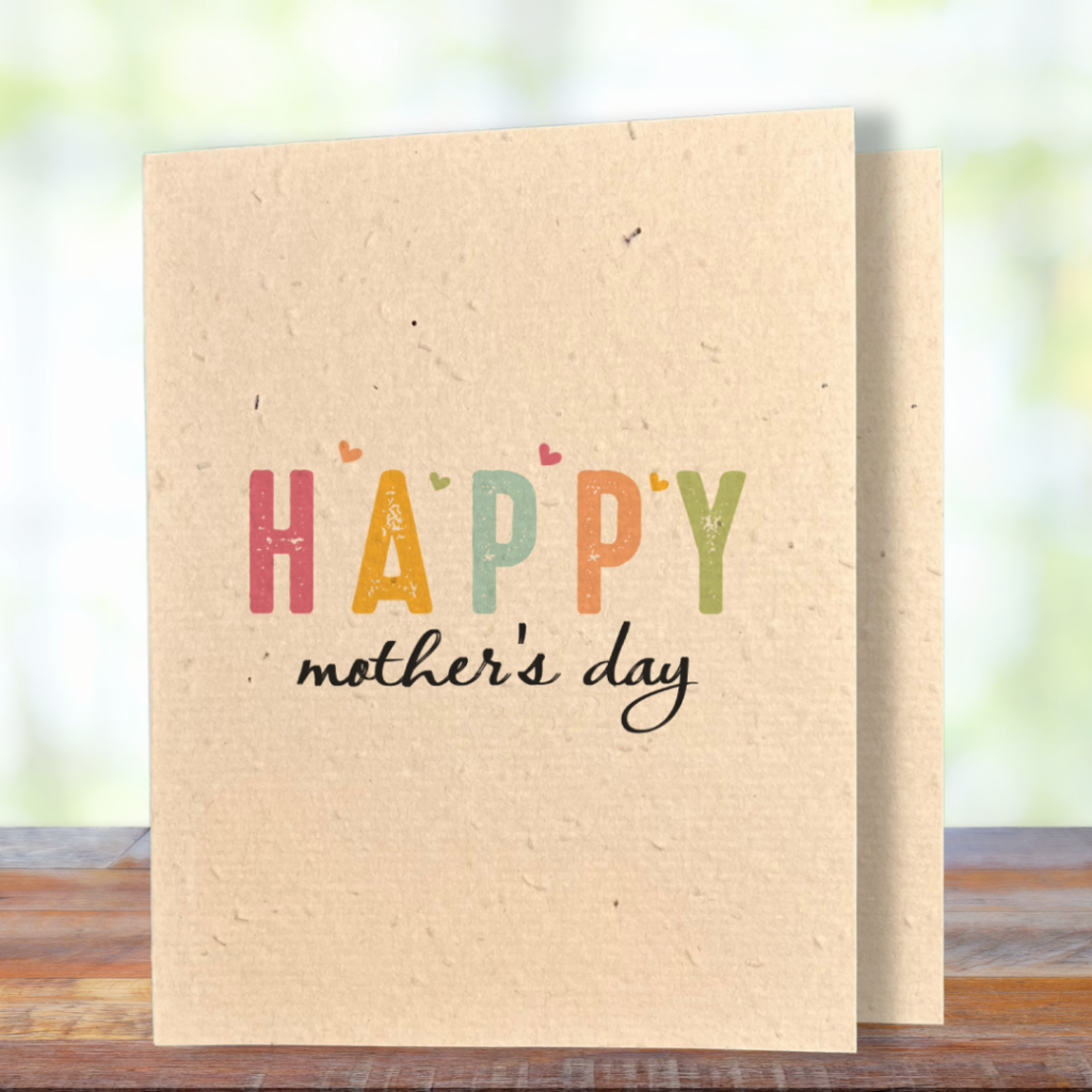 A greeting card printed on textured kraft coloured seed paper, standing upright on a wooden surface against a soft, blurred green background. The card features the word "HAPPY" in large, distressed block letters, each letter in a different colour — pink, golden yellow, teal, orange, and green — with small heart accents scattered above them. Below in flowing black script reads "mother's day."