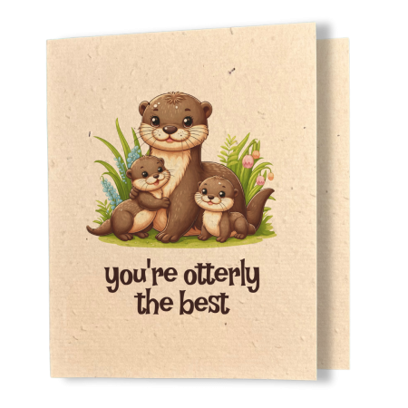 A greeting card printed on textured kraft seed paper. The card features a whimsical illustrated scene of a smiling mother otter hugging two smaller baby otters, nestled among lush green plants and delicate pink and blue flowers. Below the illustration, dark brown rounded text reads "you're otterly the best."