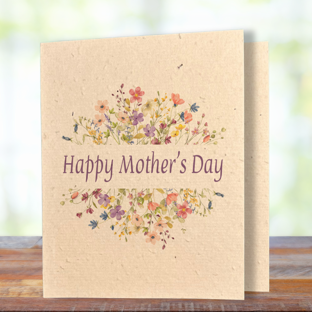 A greeting card printed on textured kraft seed paper, standing on a wooden surface against a soft blurred green background. The card features two arching clusters of delicate watercolour wildflowers in shades of pink, purple, coral, yellow, and blue, framing the text "Happy Mother's Day" in elegant purple serif lettering at the centre.
