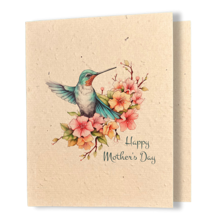 A greeting card printed on textured kraft seed paper. The card features a delicate watercolour illustration of a teal hummingbird in mid-hover, wings spread wide, resting among clusters of pink and orange blossoms on flowering branches. Below in matching teal script reads "Happy Mother's Day."